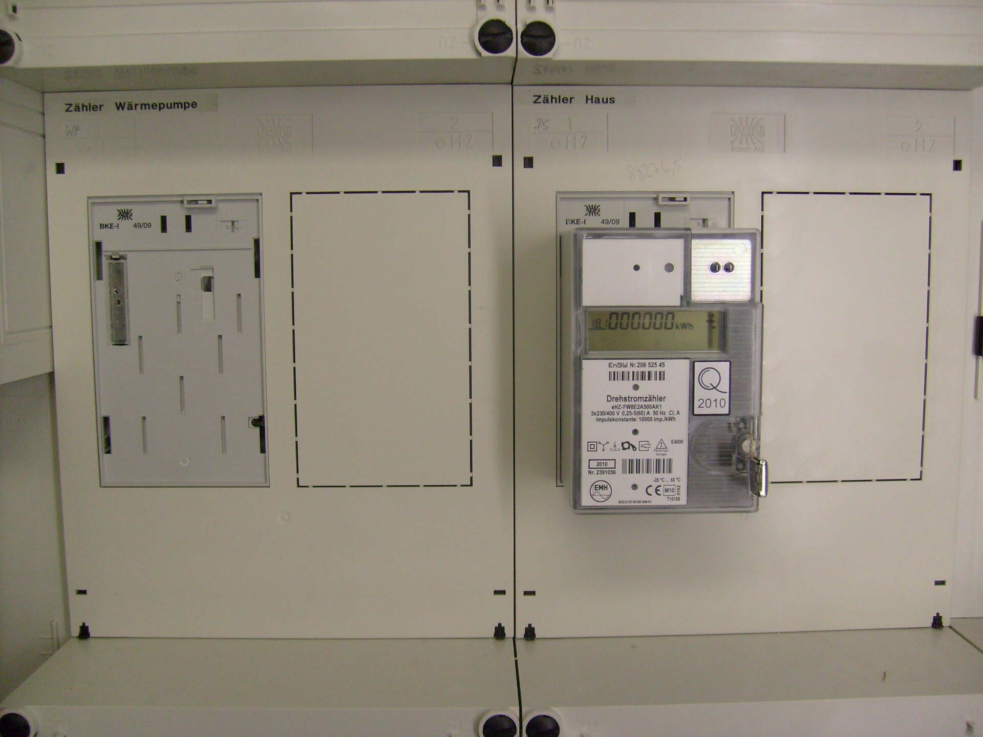 Zählerschrank mit zwei Zählerfeldern, beschriftet Zähler Wärmepumpe und Zähler Haus. Im rechten Feld steckt ein elektronischer Drehstromzähler auf einer Befestigungs- und Kontaktiereinheit, links ist die Einheit leer. Neben jedem Zähler eine gestrichelte Ausbruchfläche.